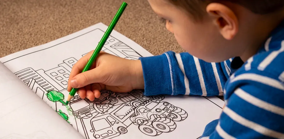A boy lying on the floor coloring a truck page green, most of the trucks still outlines