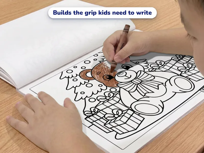 Child with a crayon coloring a picture of a teddy bear sitting in front of a decorated xmas tree